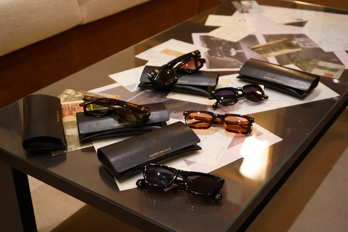 Les cinq lunettes de soleil Maison Melville en acétate noir et écaille, avec leurs étuis noirs, sur une table couverte de photographies anciennes